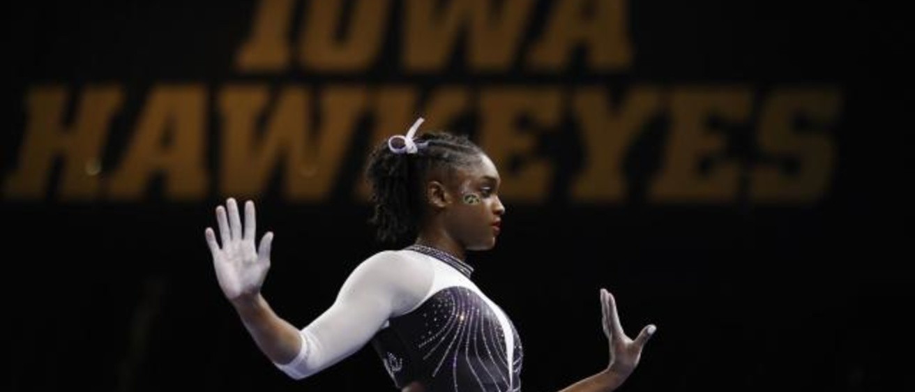An gymnast stands with her arms and hands out to her sides against a black wall with the words "Iowa Hawkeyes" in gold.